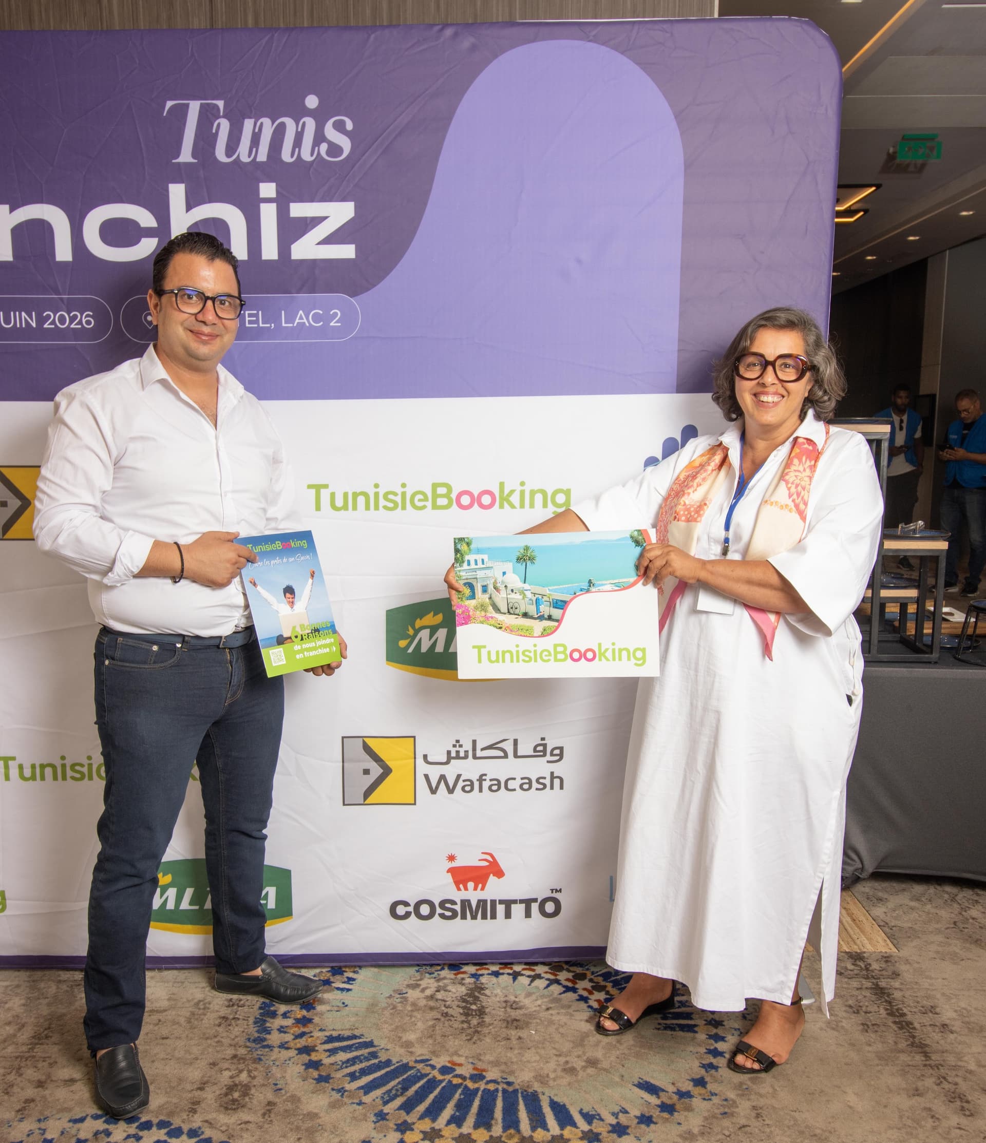 ممثل TunisieBooking ومنظمة الحدث يحملان صورًا ترويجية للعلامة التجارية أمام منطقة التصوير في WeFranchiz Days Tunis 2026.